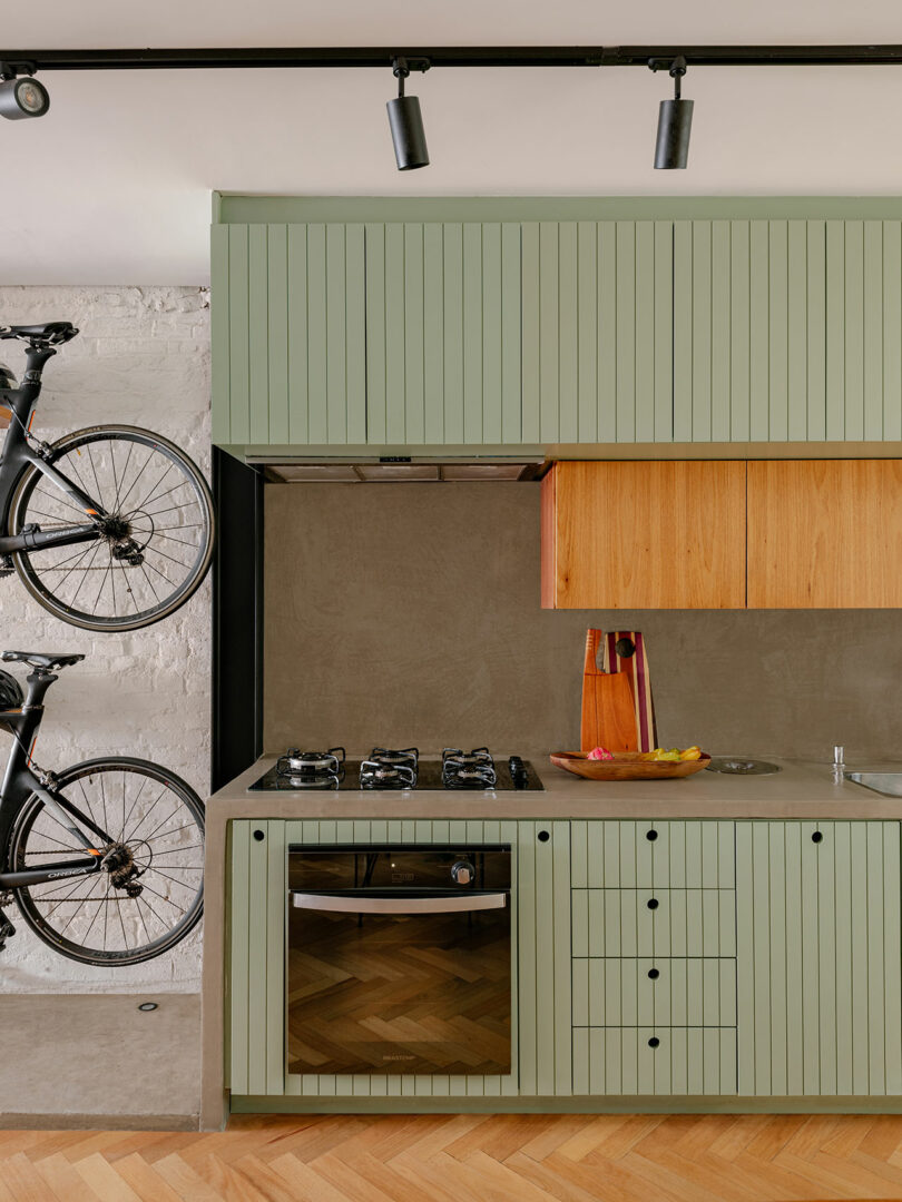 Modern kitchen with sage green cabinets, wooden upper cabinets, built-in oven, gas stove, and two bicycles mounted vertically on the adjacent wall.