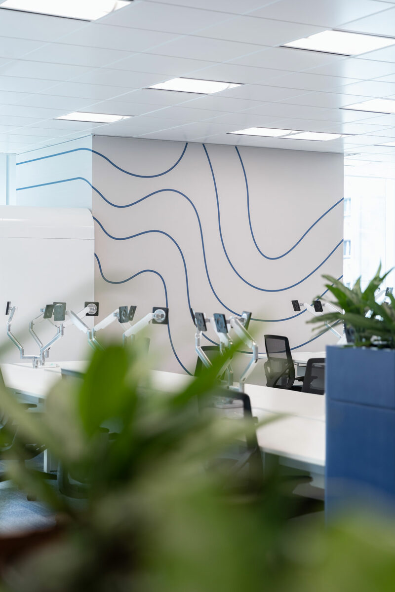 Modern office with empty desks, computer monitors on adjustable arms, black chairs, and a white wall featuring abstract blue curved lines; green plants in the foreground.
