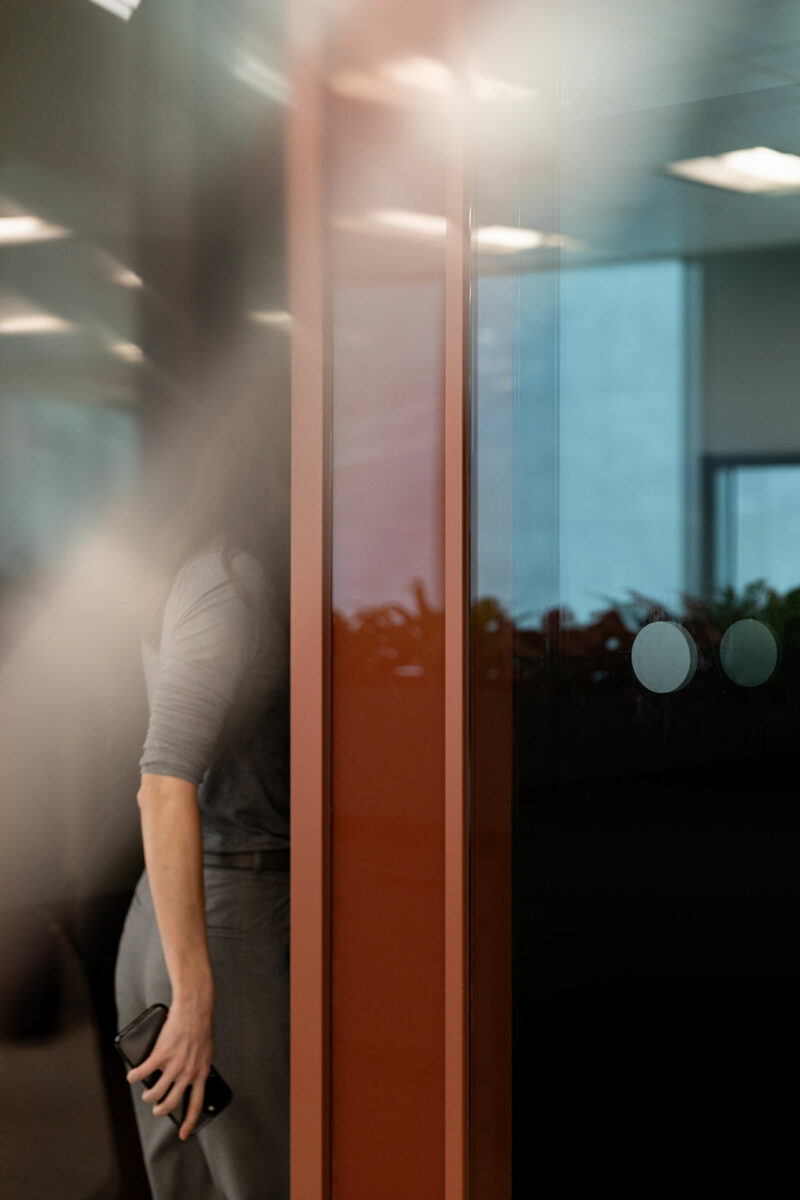 A person in gray clothing, partially obscured by a glass wall, stands near a red frame while holding a smartphone in their right hand.
