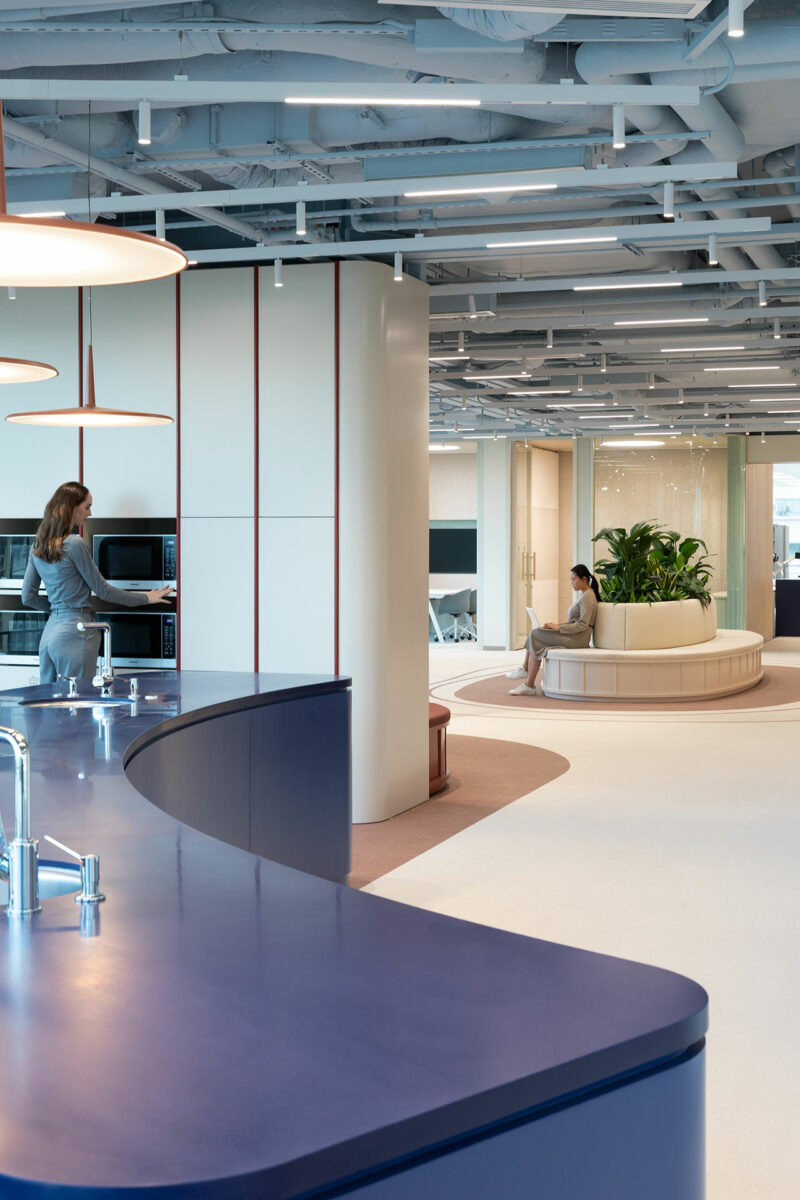 Modern office interior with a curved blue counter, a woman using a kitchen area, and another person seated on a circular bench near a plant.