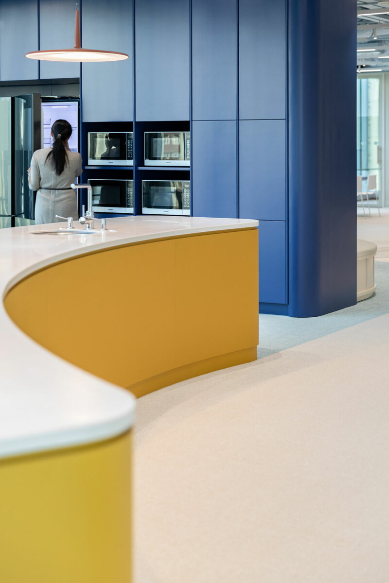 A person stands at a refrigerator in a modern kitchen with blue cabinets, built-in microwaves, and curved yellow counters.