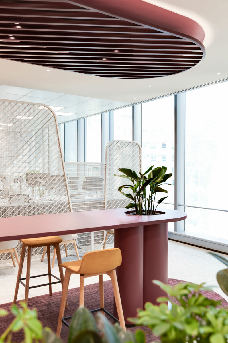 Modern office space with a pink table, wooden stools, a potted plant, large windows, and decorative room dividers.