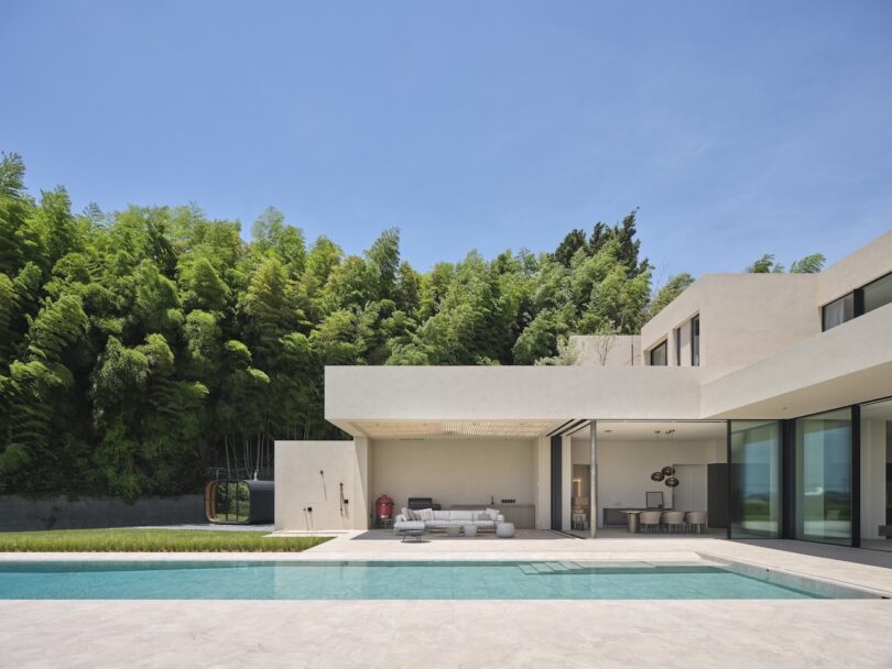 Modern house with large windows, outdoor lounge area, and a swimming pool in front, surrounded by lush green trees and clear blue sky.