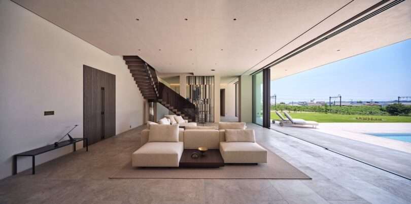 Modern living room with beige sofas, a wooden coffee table, and an open wall leading to a patio, pool, and outdoor lawn with lounge chairs and a view of the ocean.