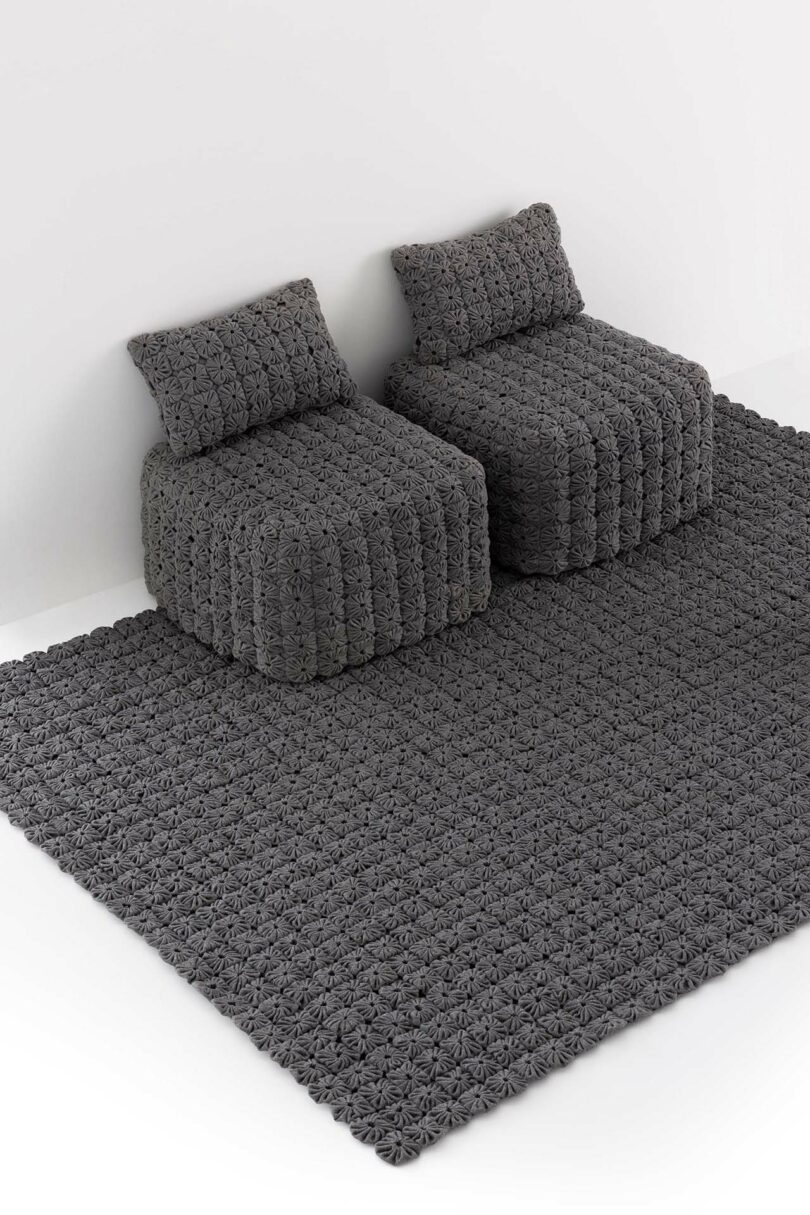 Two chunky, textured gray armchairs with matching pillows are positioned on a large, patterned gray rug against a white wall.