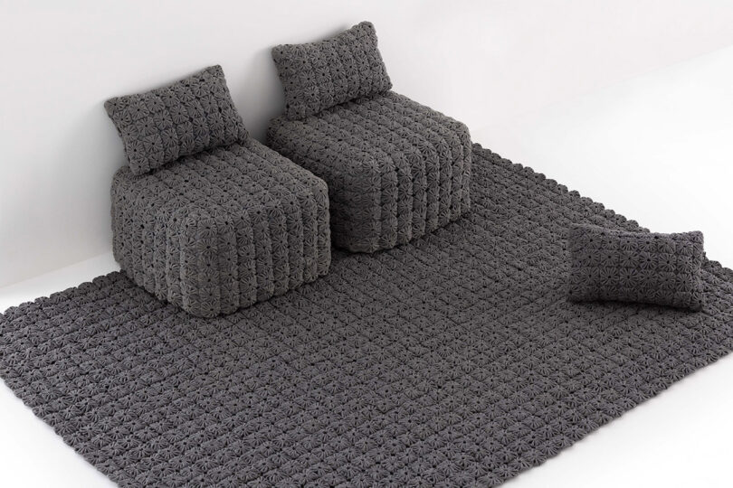 Two textured gray armchairs and a pillow sit on a matching textured gray rug in a minimal, white room.