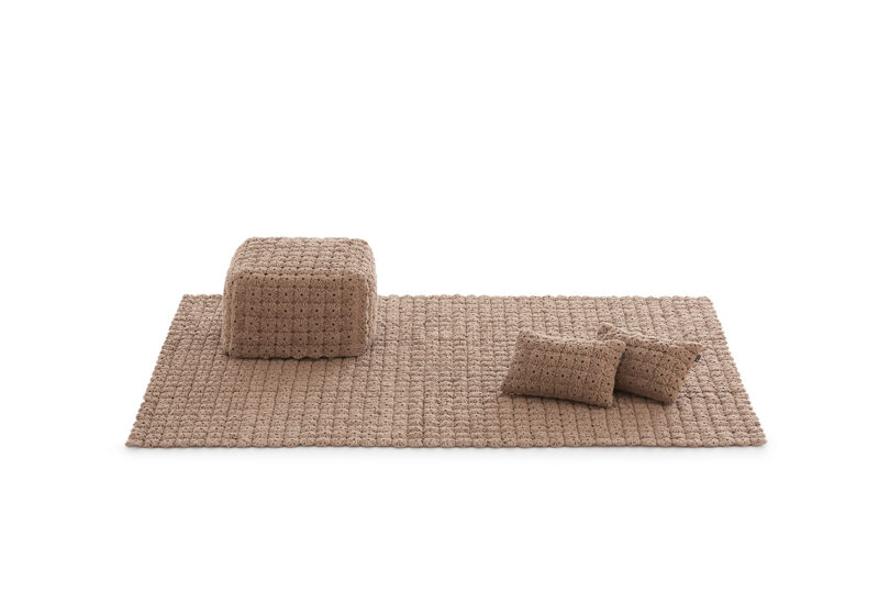 A beige woven rug with two matching pillows and a cube-shaped pouf, all set against a white background.