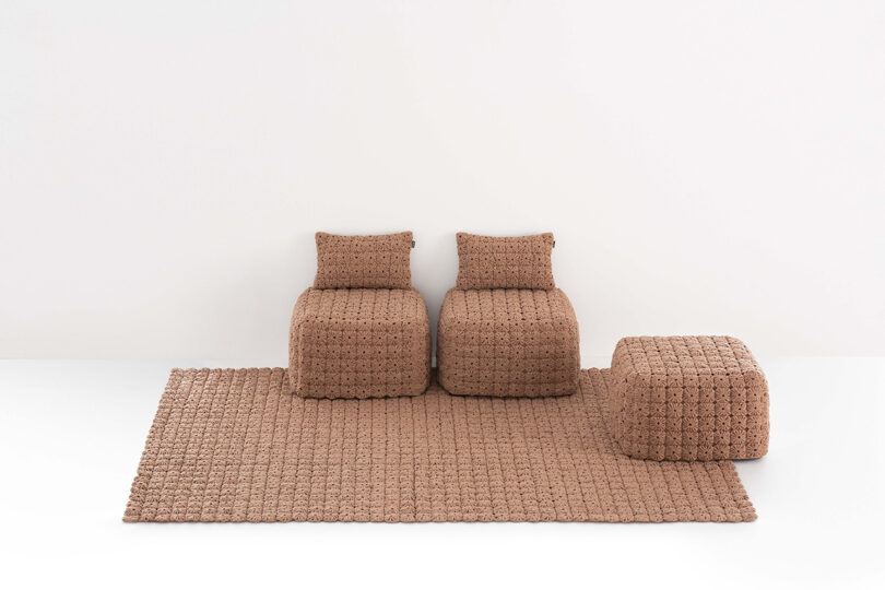 Two brown textured armchairs with matching cushions are placed on a square brown textured rug, alongside a matching ottoman, against a plain white background.