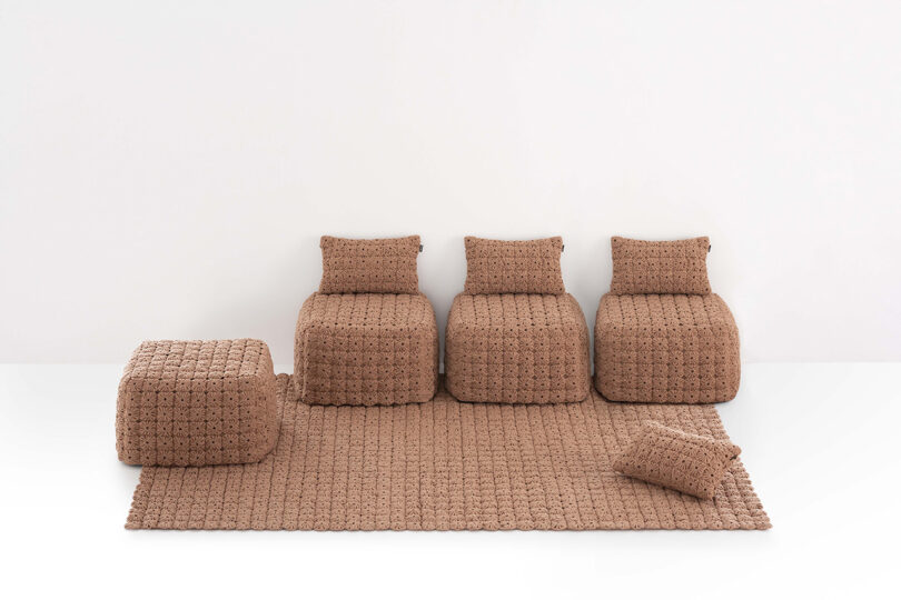 Three brown textured chairs, a matching ottoman, a cushion, and a large rug are arranged against a plain white background.