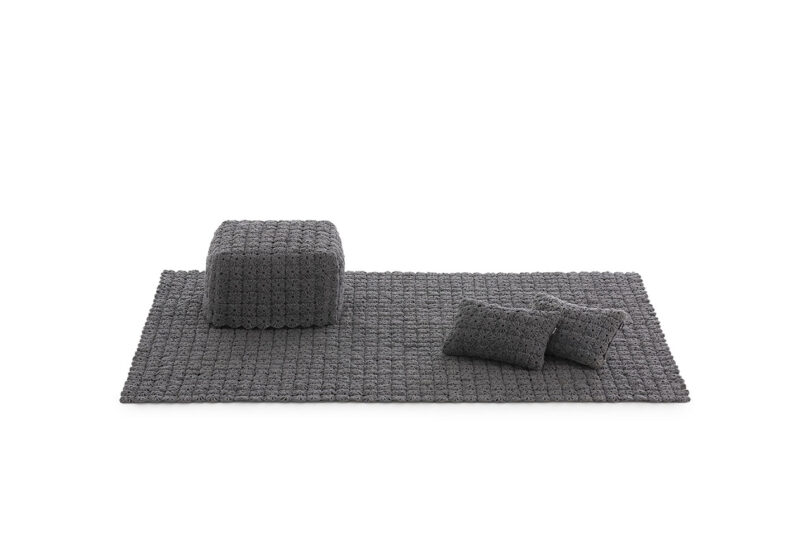 A textured gray rug with a matching square pouf and two rectangular pillows, all in a similar knitted pattern, set against a white background.