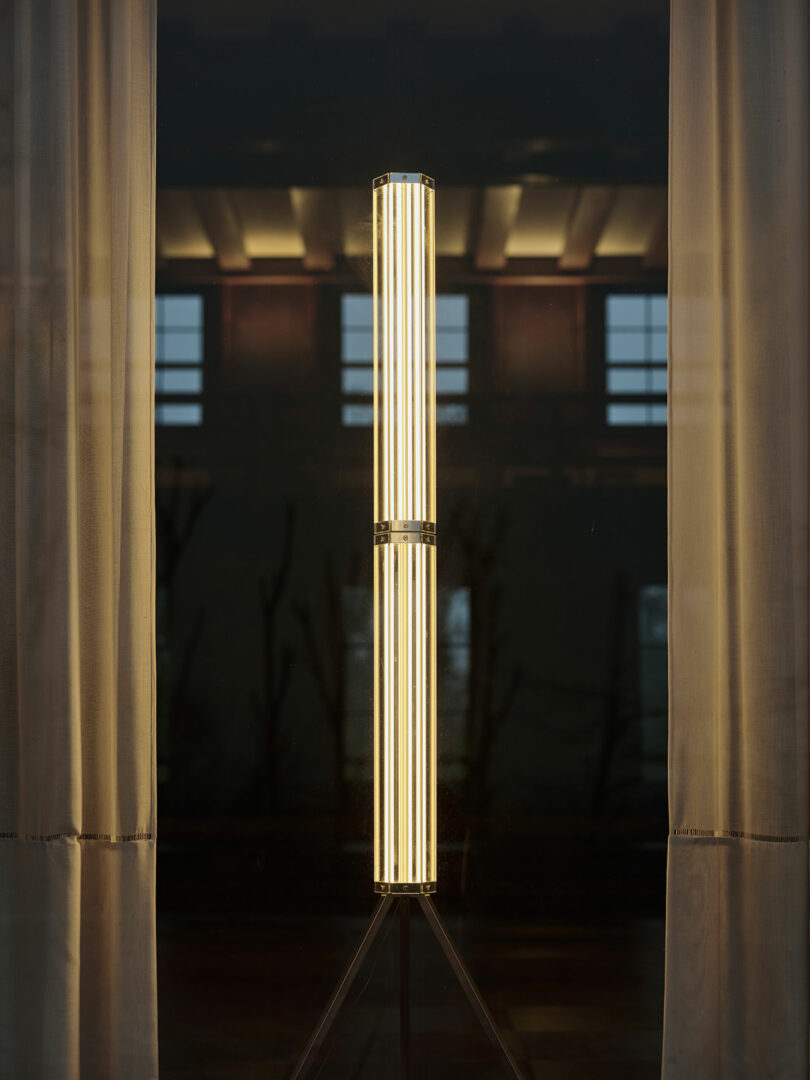 A vertical cylindrical lamp with multiple LED tubes emits warm light, standing between two beige curtains in front of a window with a dark background.