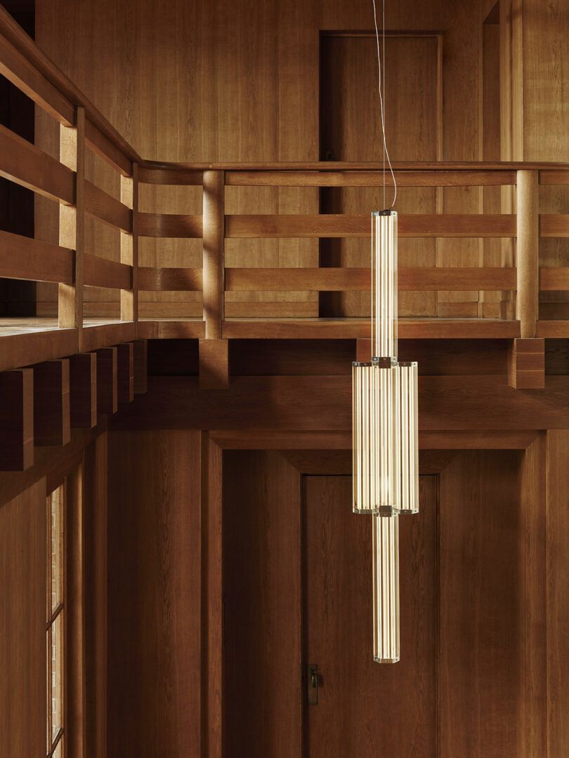 A modern, vertical pendant light with cylindrical bulbs hangs in a two-story interior featuring wooden walls, railings, and trim.