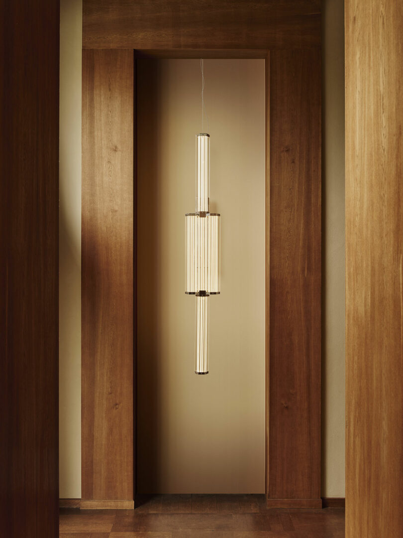 A modern, cylindrical pendant light with vertical slats hangs in a wooden hallway with warm, neutral tones.