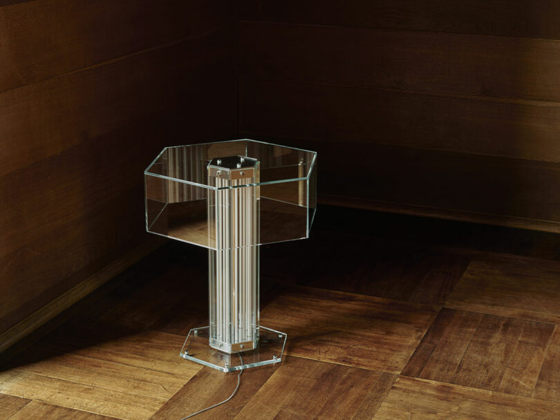 A transparent hexagonal acrylic side table with a column base stands on a wooden floor in the corner of a room with wood-paneled walls.