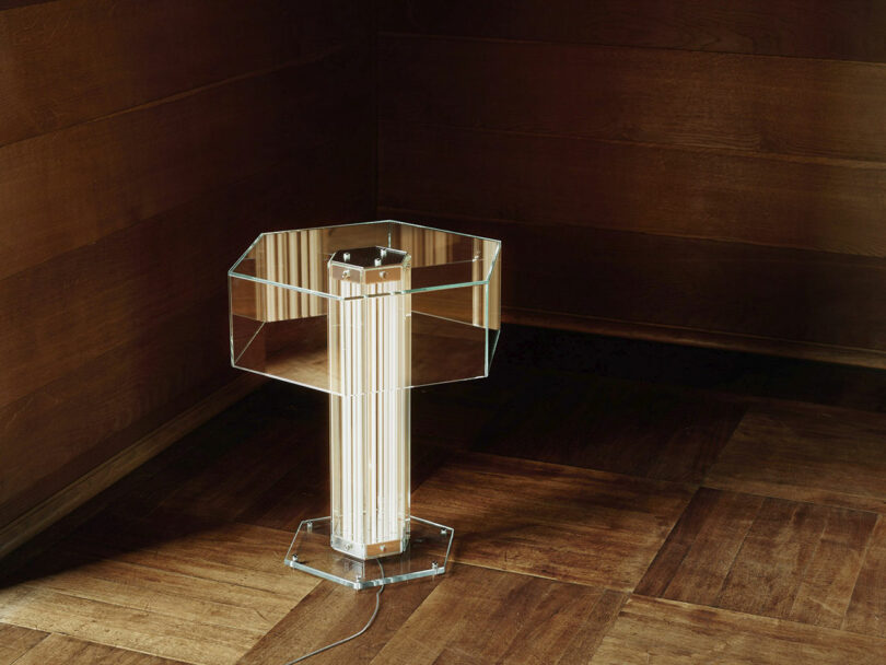 A clear hexagonal pedestal lamp with vertical light tubes stands on a wooden floor in the corner of a room with wooden-paneled walls.