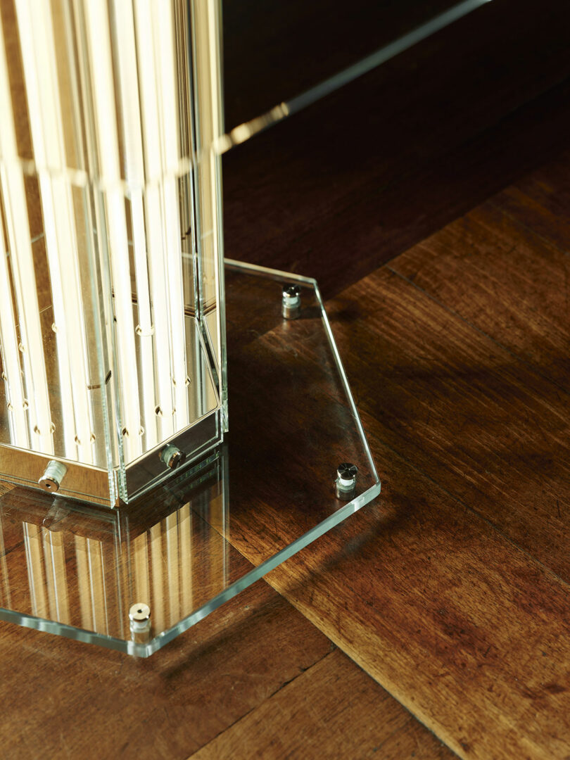 A close-up of a glass hexagonal base with metal bolts, supporting a vertical glass or acrylic structure on a wooden floor.
