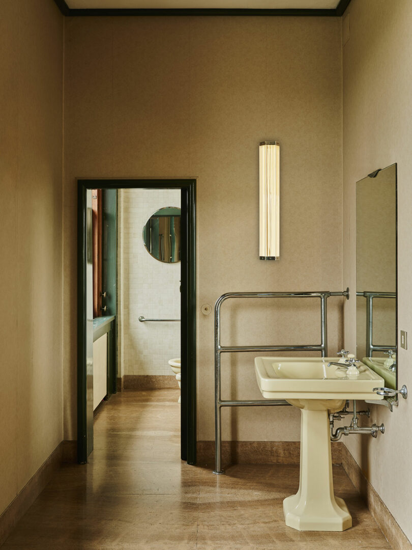 A beige bathroom with a pedestal sink, wall mirror, metal towel rail, and vertical light fixture; an open door reveals a second bathroom with a round mirror and toilet.