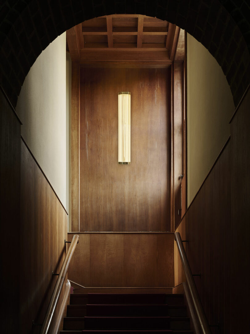 A wooden staircase with paneled walls leads up to a landing with a rectangular wall sconce emitting warm light, framed by an arched brick entryway.