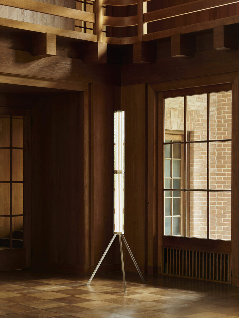 A tall, cylindrical floor lamp with a tripod base stands in the corner of a wood-paneled room with large windows and wooden architectural details.
