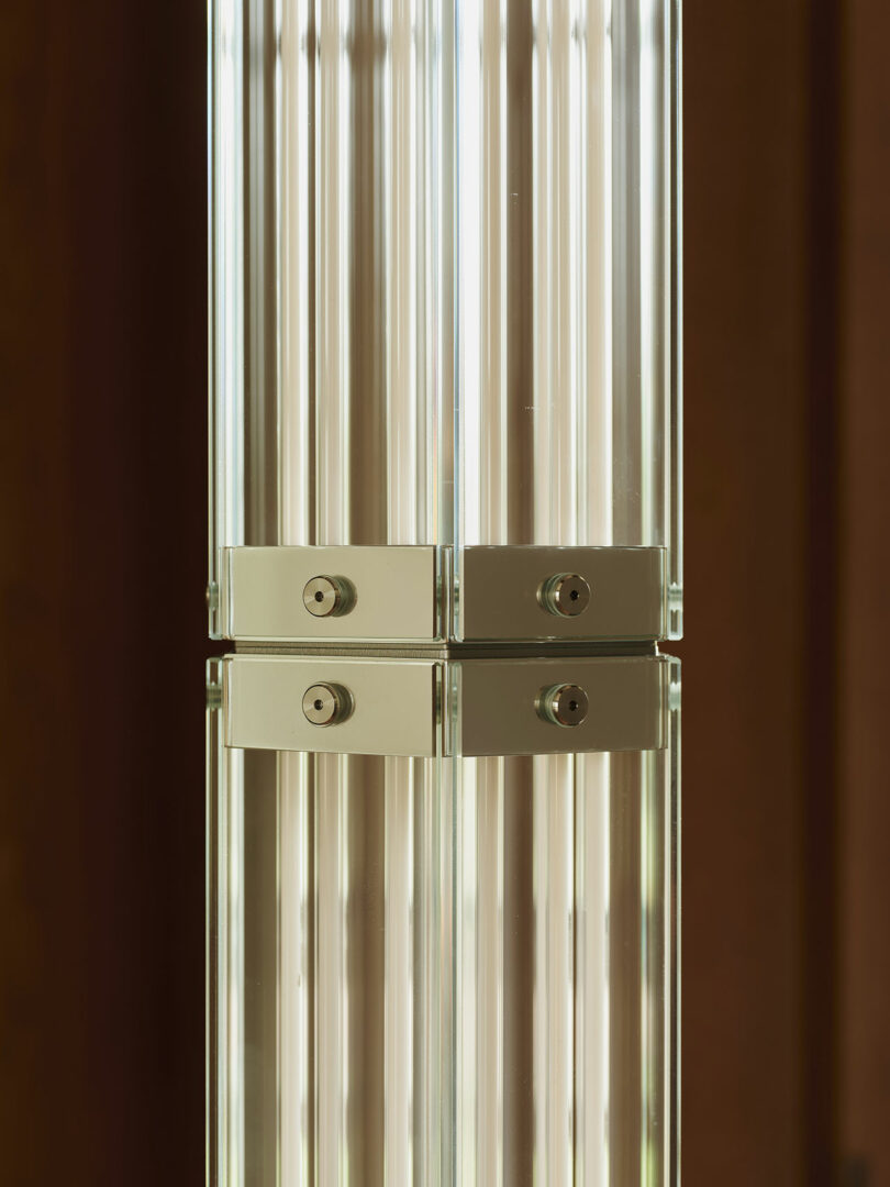 Close-up of a reflective metallic and glass column with vertical lines and two metal connectors, set against a blurred brown background.