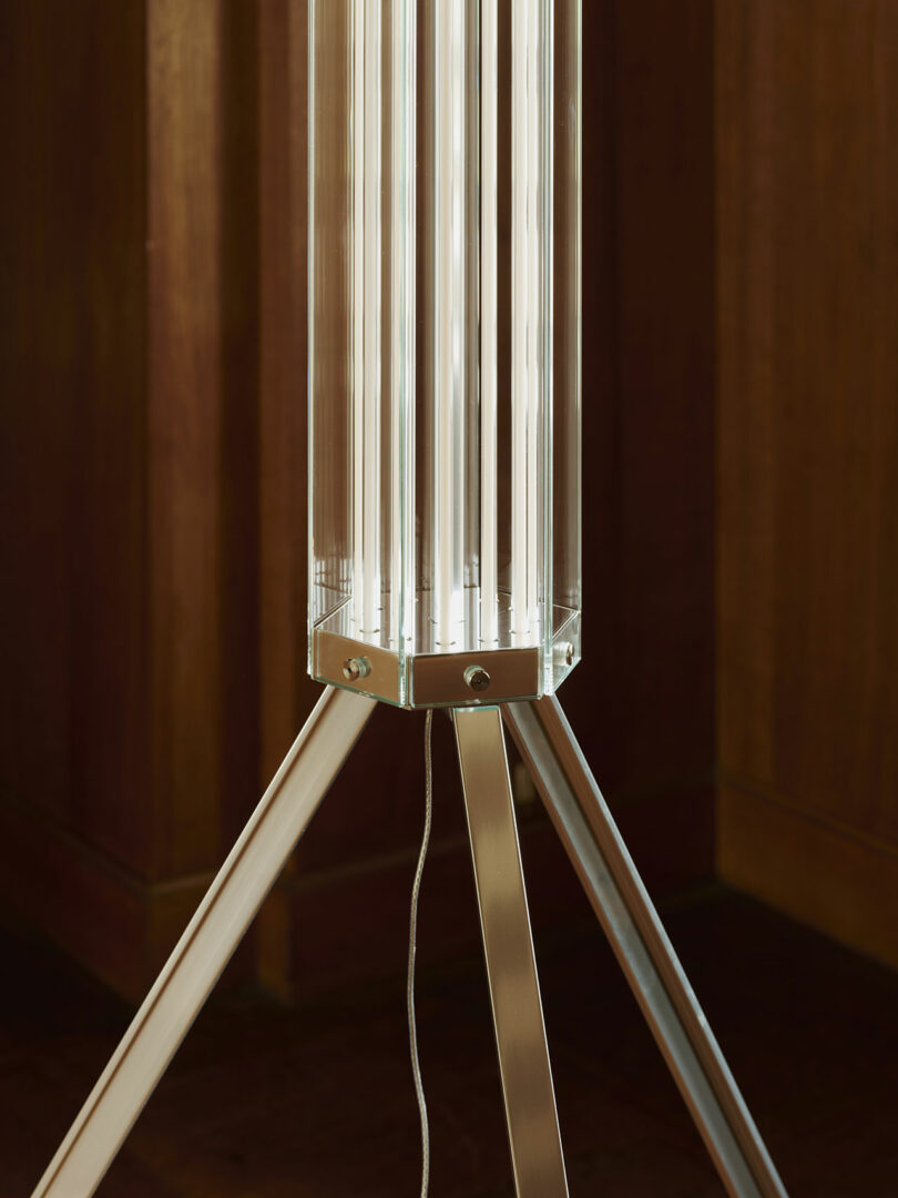 A modern floor lamp with vertical glass tubes on a metal tripod base, positioned against a wooden wall.