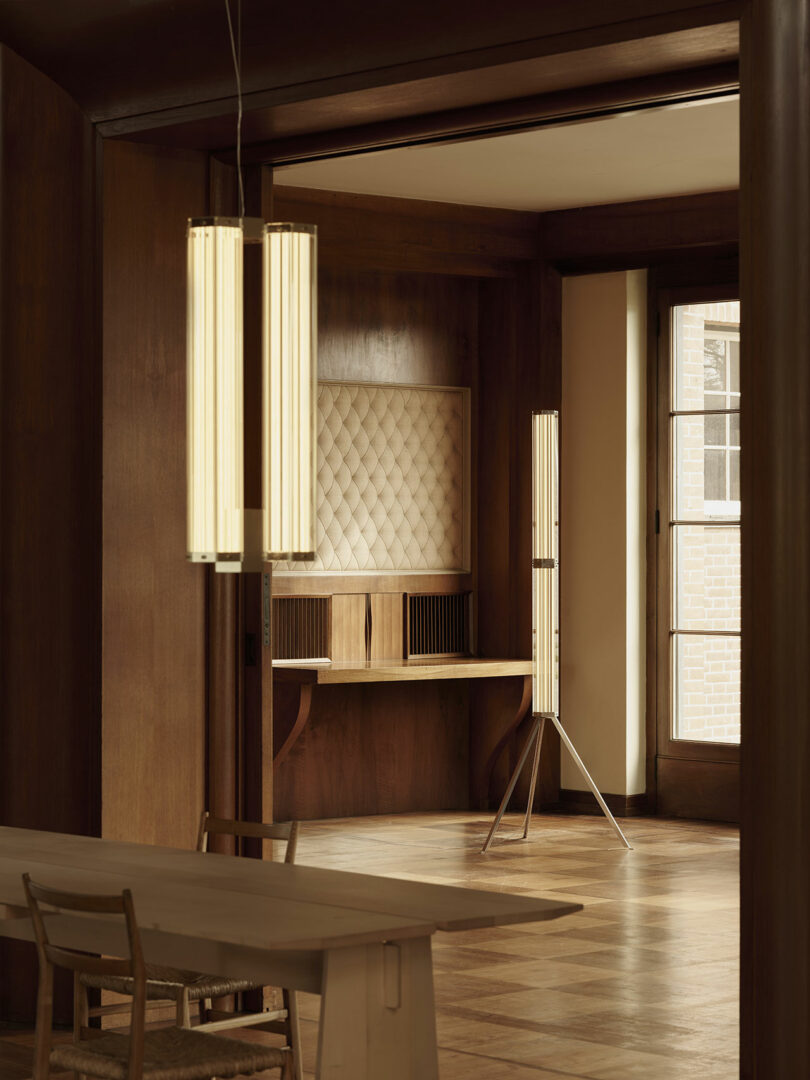 A minimalist room features wood-paneled walls, a built-in desk with a quilted back panel, modern vertical light fixtures, and a large window letting in natural light.