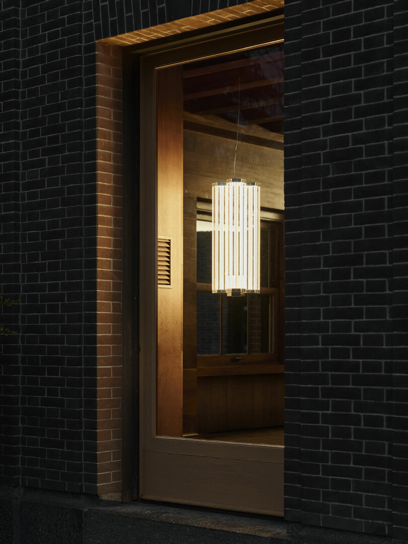 A cylindrical pendant light hangs inside a room with dark brick walls, seen through a large glass window at night.
