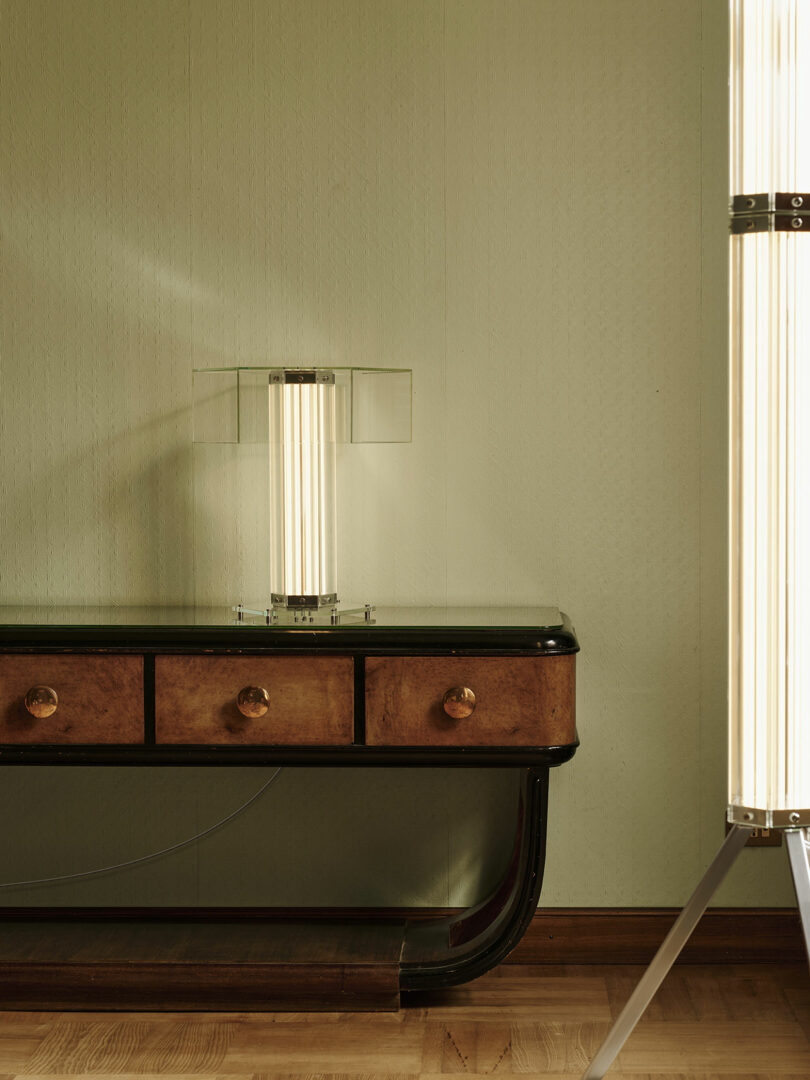 A glass and wood console table with three drawers and round knobs stands against a light green wall, with a modern vertical lamp on top and another similar lamp to the right.