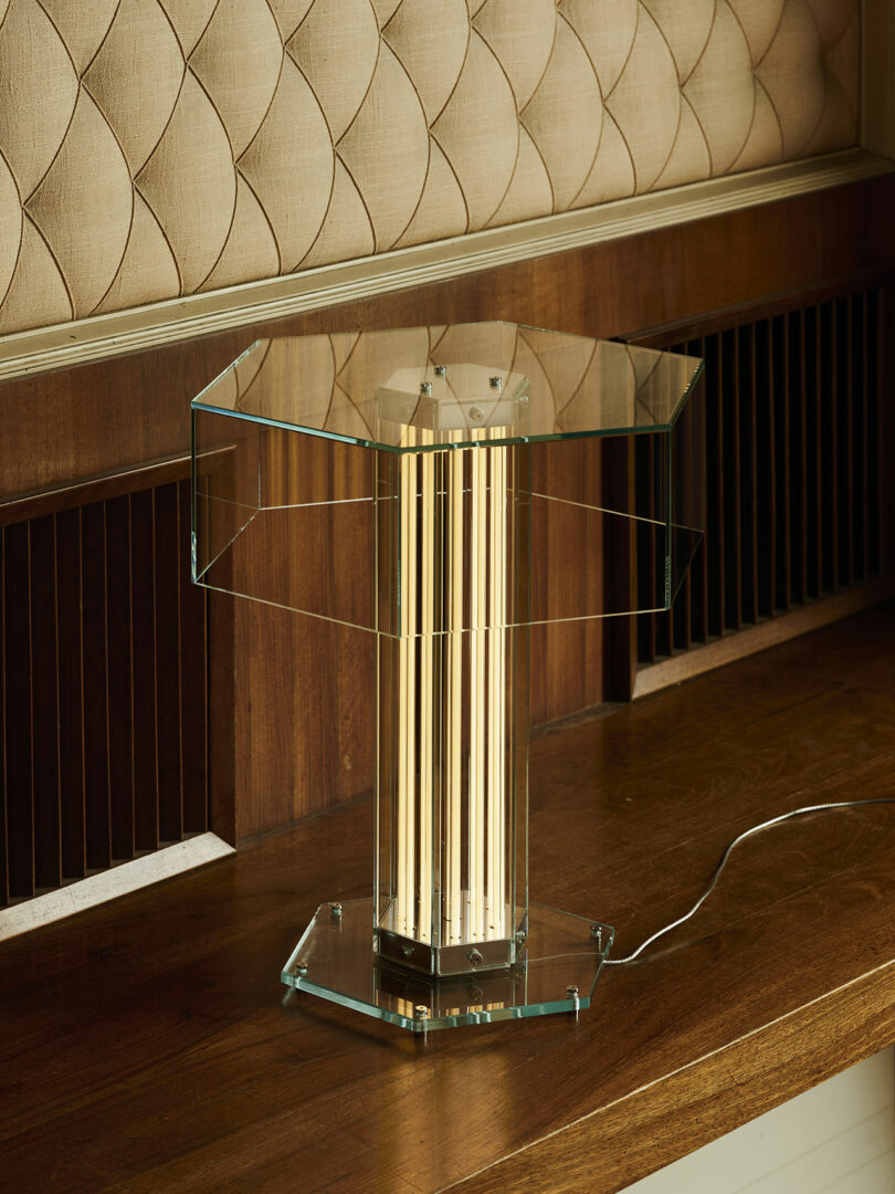 A modern glass table lamp with vertical light tubes stands on a wooden surface, with its power cord visible against a wood and fabric-paneled wall.