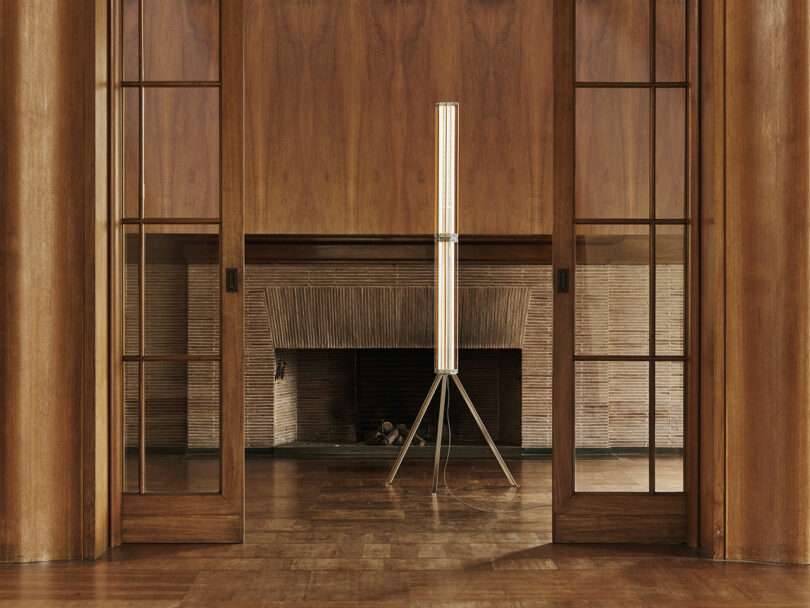 A modern floor lamp with a cylindrical shade stands in front of a fireplace, surrounded by wooden paneling and sliding glass doors in a spacious room.