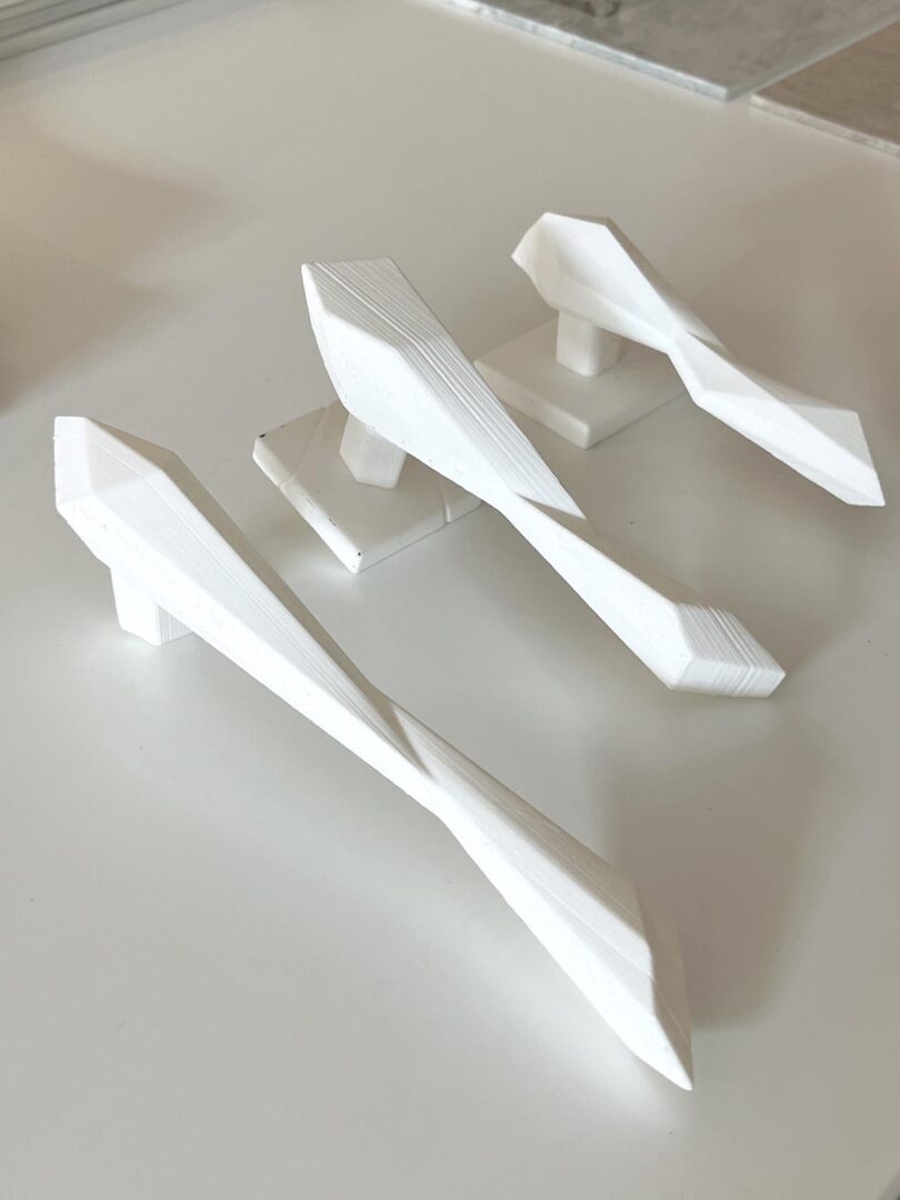 Three white geometric, abstract bird-like sculptures are displayed on flat bases atop a light-colored surface.