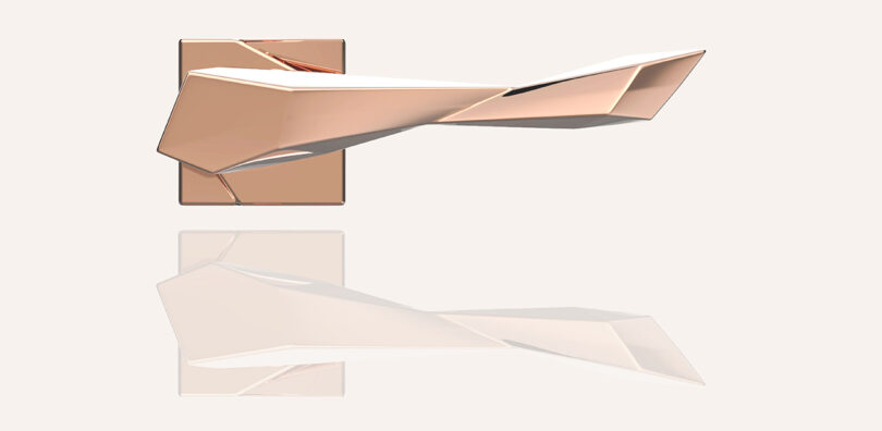 Angular, rose gold door handle with a geometric design mounted on a matching square plate, reflected on a glossy surface.