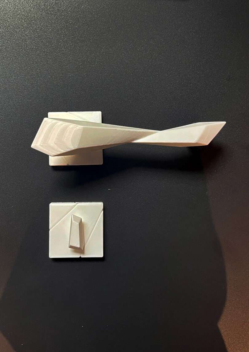 A white, angular door handle and its matching square backplate placed on a black surface.