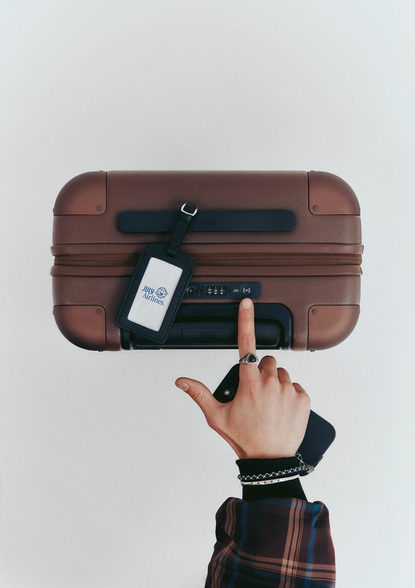A person holding a brown suitcase by the handle, with a luggage tag attached and pointing at the suitcase lock, holding a phone in the other hand.