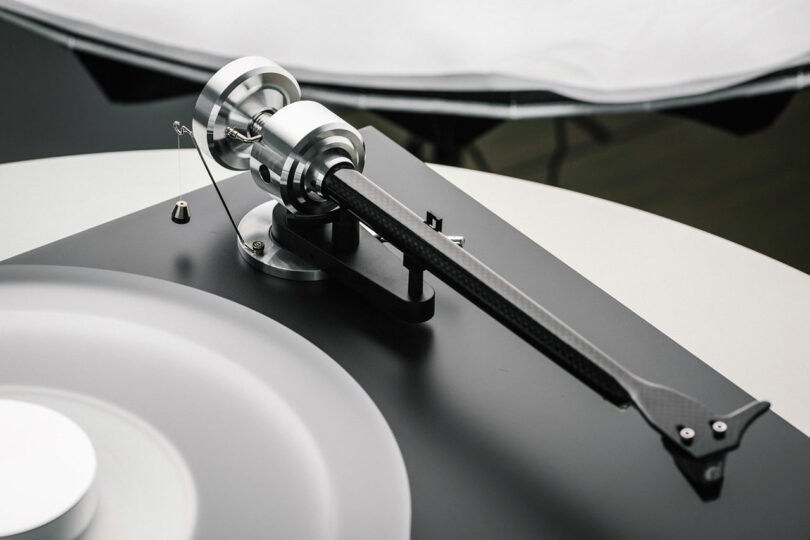 Close-up of a modern turntable tonearm and cartridge mounted on a black base, with metallic and carbon fiber elements visible.