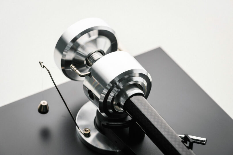 Close-up of a high-precision metallic tonearm mechanism on a turntable, featuring a counterweight and an attached thread with a small weight.