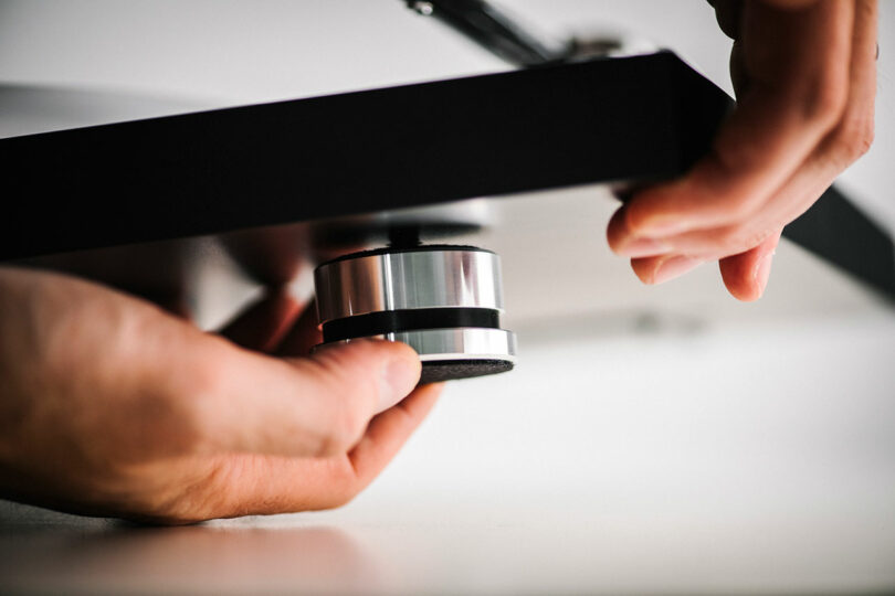 Close-up of hands adjusting a cylindrical metal leveling foot attached to the underside of a black flat surface, possibly furniture or equipment.