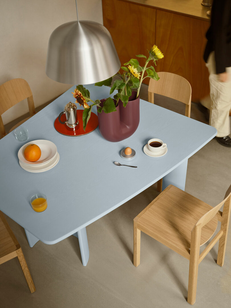 A light blue table set with a vase of sunflowers, a cup of coffee, an egg, fruit, a glass of orange juice, and a metal coffee pot under a silver pendant lamp.