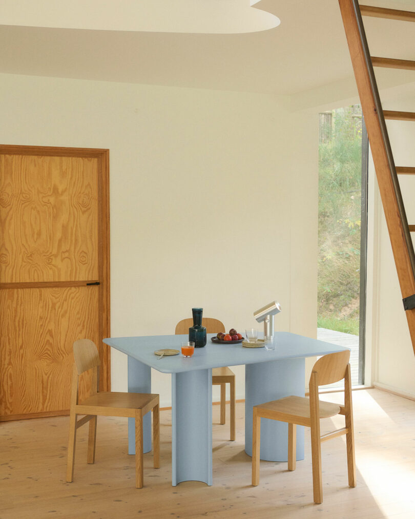 A minimalist dining area with a light blue table, three wooden chairs, assorted tableware and fruit, a wooden door, a ladder, and large window with greenery outside.