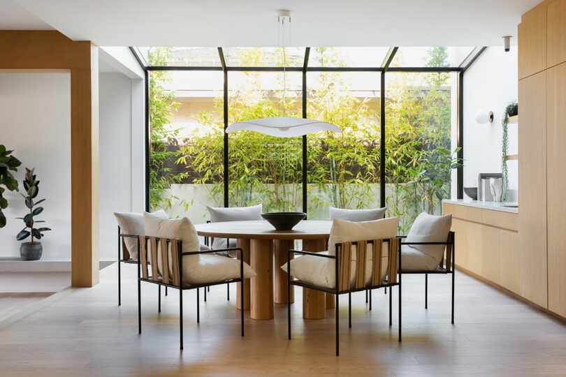 A modern dining room with a round wooden table, six cushioned chairs, and a large window overlooking bamboo plants.