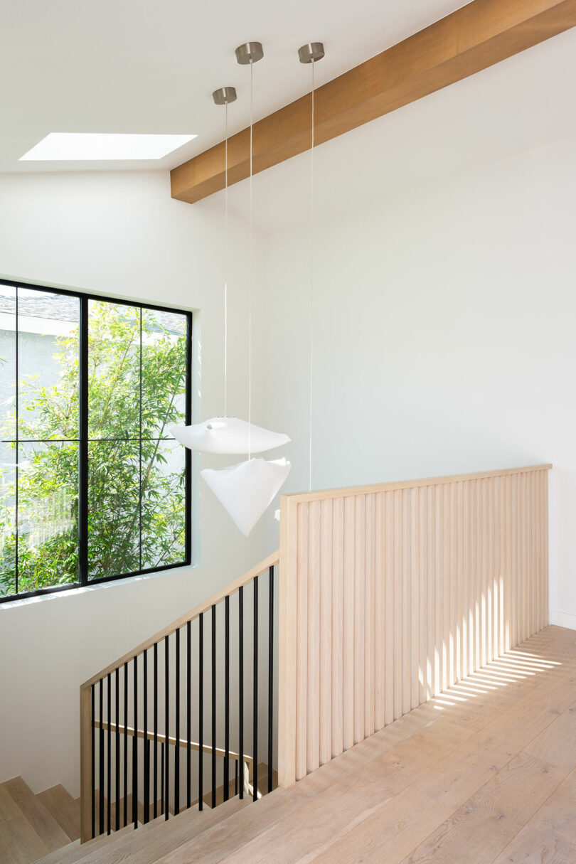 Modern stairway with light wood railing, vertical black balusters, large window with outdoor greenery, and two white geometric pendant lights hanging from the ceiling.