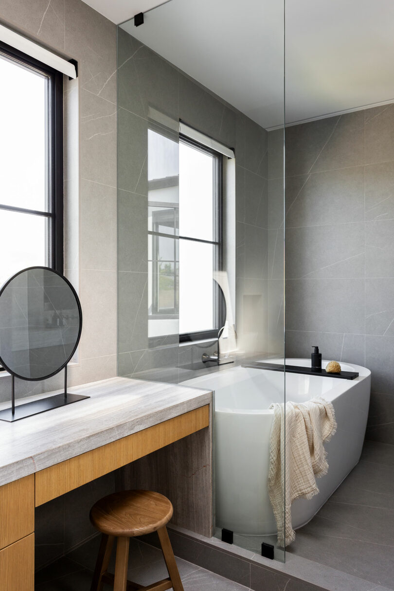 Modern bathroom with a freestanding bathtub, a draped towel, a wooden vanity with a round mirror, a wooden stool, and large windows letting in natural light.