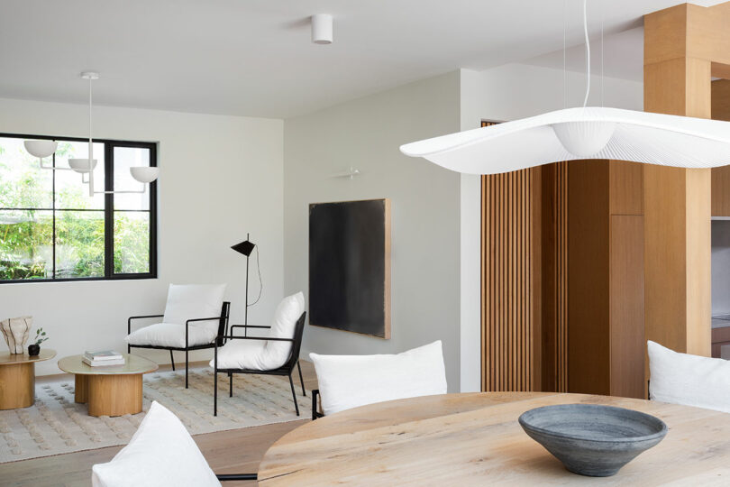 Modern living and dining area with light wood furniture, white cushions, a round dining table, large window, minimalist decor, and neutral color palette.