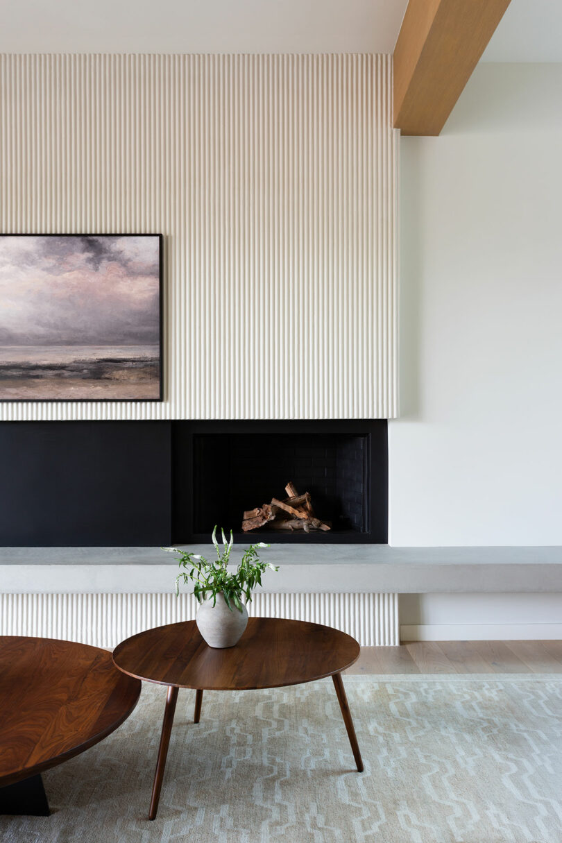 A modern living room with a wood coffee table, a potted plant, a fireplace with stacked logs, and a landscape painting above the mantel.