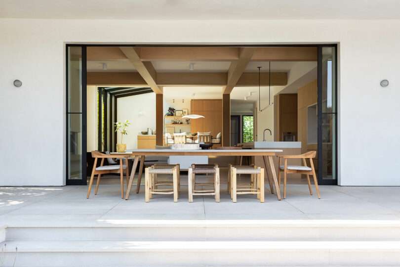 Modern open-plan kitchen and dining area viewed from a patio with a large wooden table and chairs, beige tile flooring, and minimalist décor.
