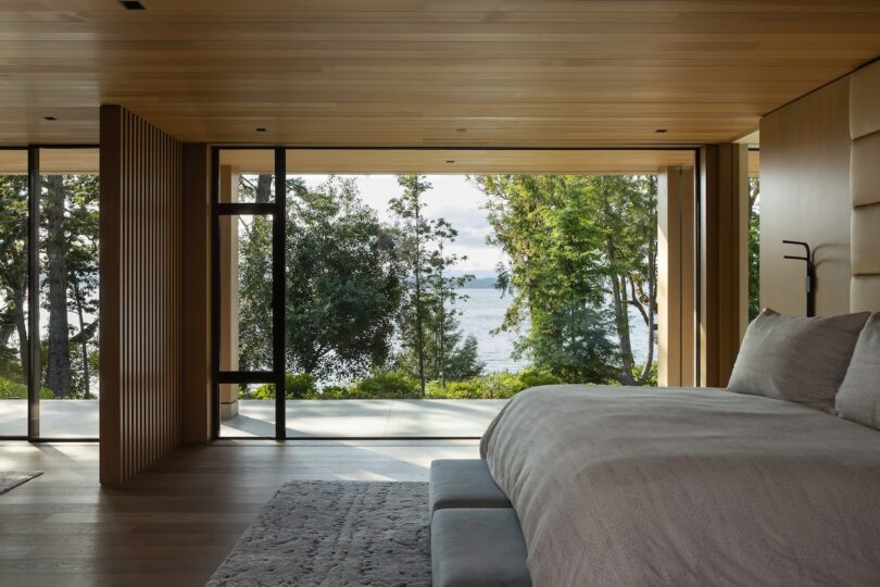 Modern bedroom with large windows and wooden walls overlooking trees and a body of water, featuring a neatly made bed and minimalist decor.