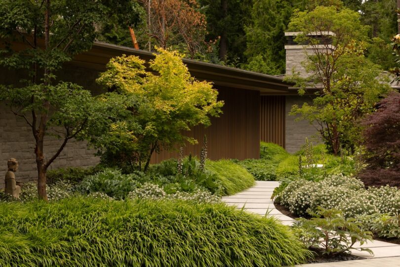 A modern house with stone walls and a slatted facade, surrounded by lush green landscaping and a curved pathway.