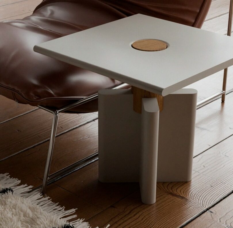 A modern square side table with a circular wooden accent stands next to a brown leather lounge chair on wooden flooring with a white rug nearby