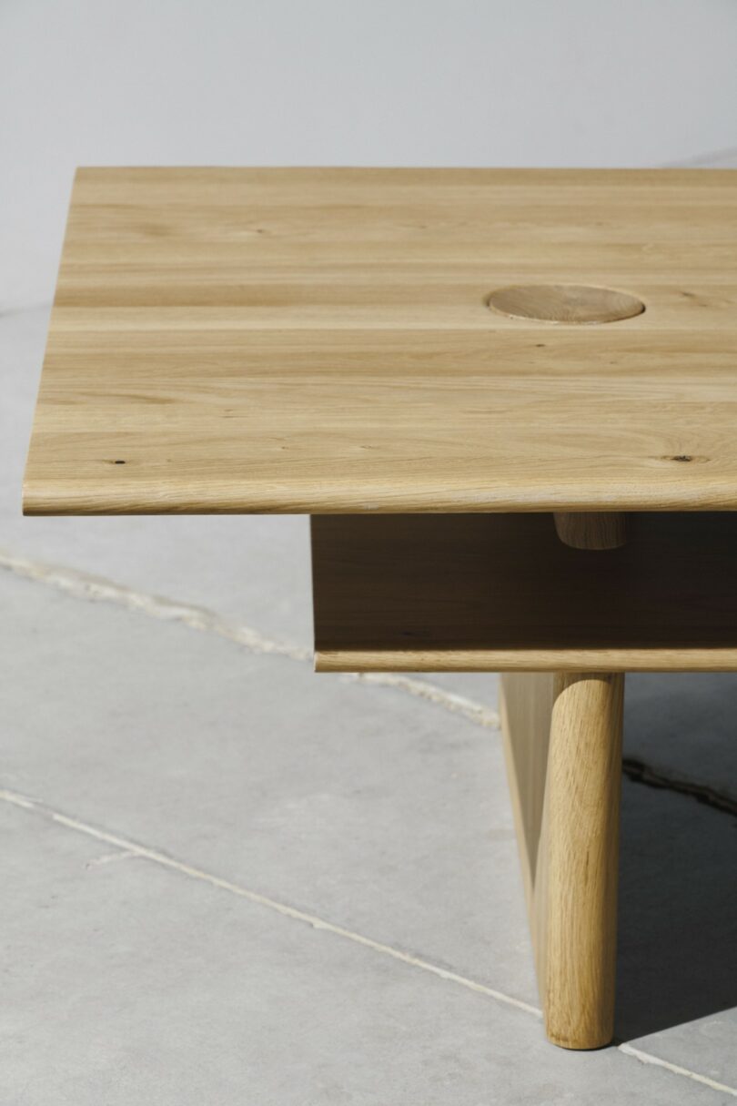 A minimalist wooden table with a rectangular top, a circular indentation, and simple, sturdy legs, placed on a light stone floor