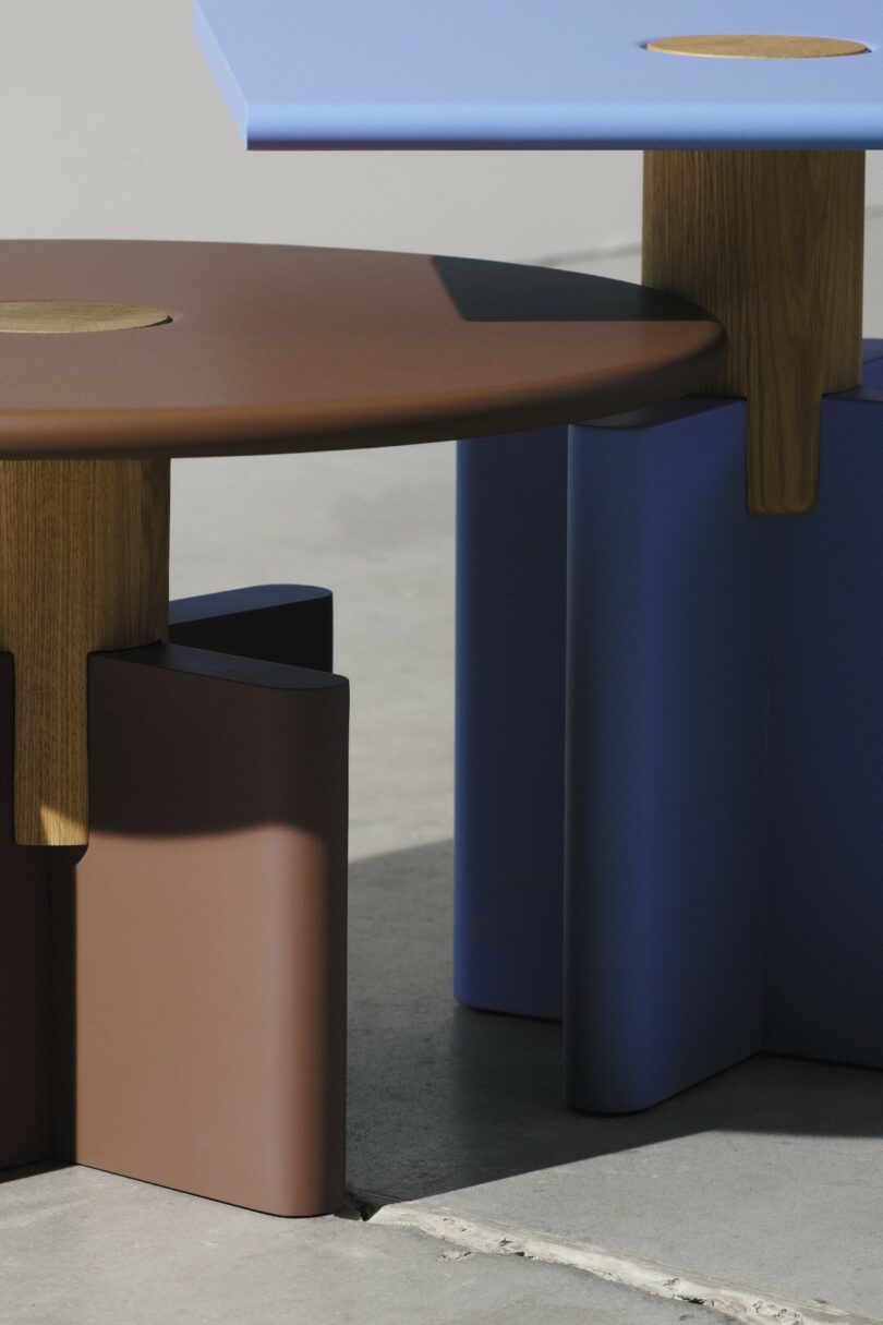 Two modern tables with rounded edges and wooden legs; one table is brown, the other is blue, both standing on a concrete floor